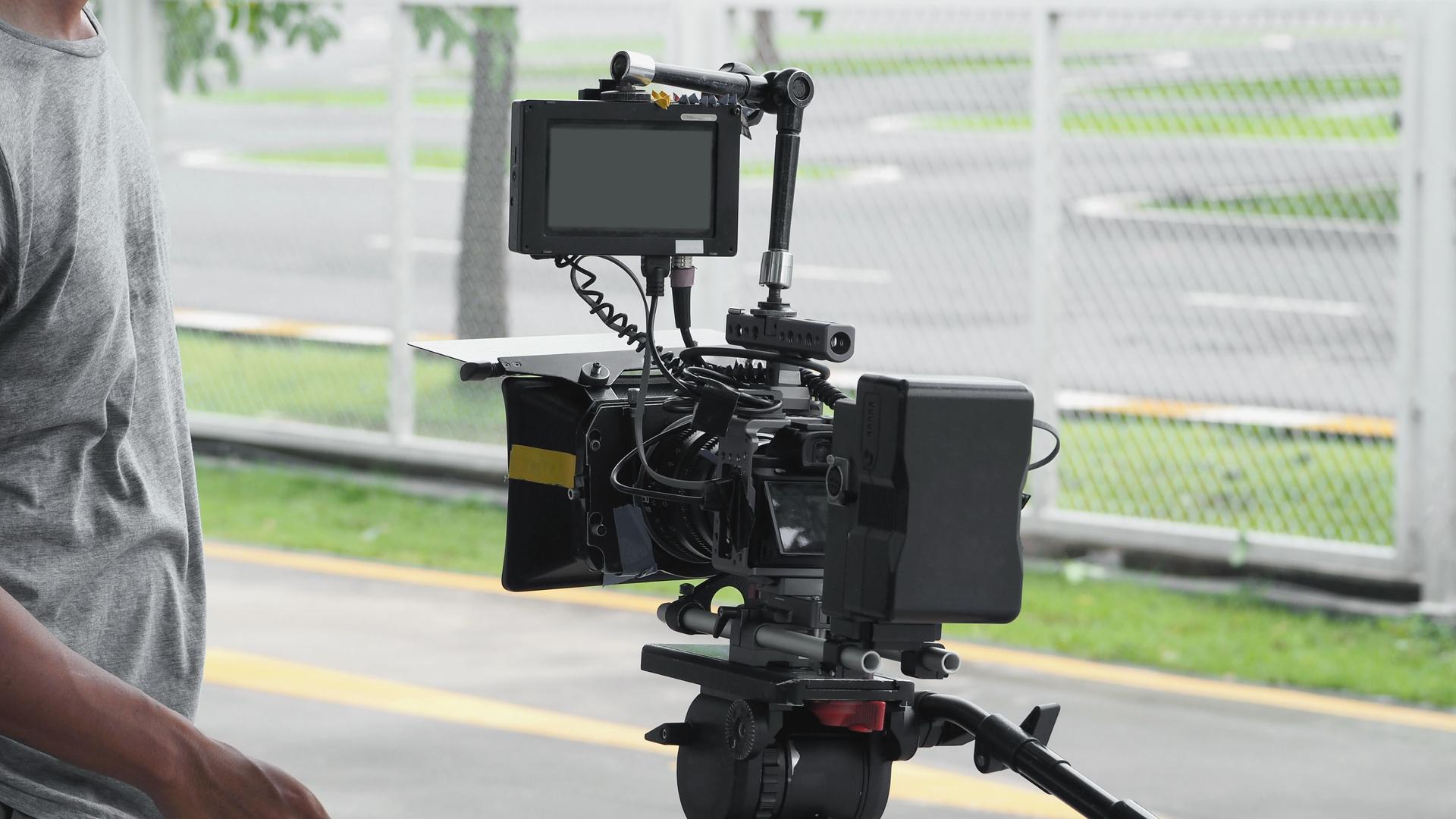Back angle view of high definition digital video camera which recording or shooting tv commercial production on pan tilt shift professional head tripod at outdoor location and film crew team with set.